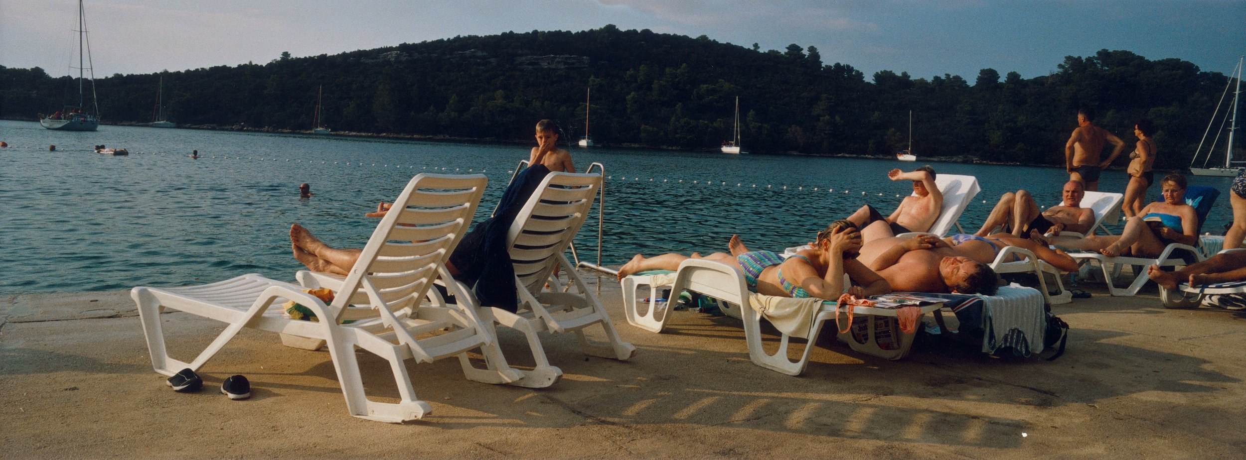 People lounge on chairs by the sea with sailboats and green hills in the background on a sunny day at a coastal resort.
