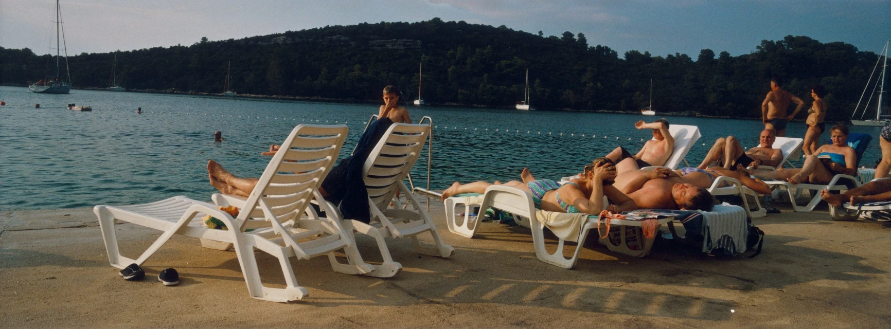People lounge on chairs by the sea with sailboats and green hills in the background on a sunny day at a coastal resort.