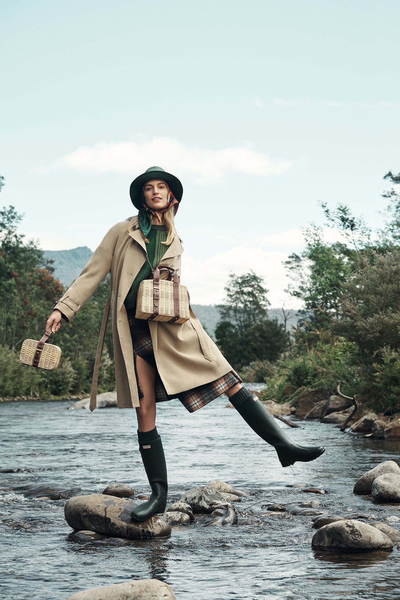 A woman in a trench coat stands on rocks in a river, posing with bags and green rubber boots against a natural, outdoor backdrop.