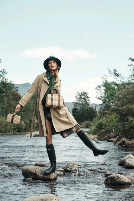 A woman in a trench coat stands on rocks in a river, posing with bags and green rubber boots against a natural, outdoor backdrop.