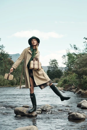 A woman in a trench coat stands on rocks in a river, posing with bags and green rubber boots against a natural, outdoor backdrop.