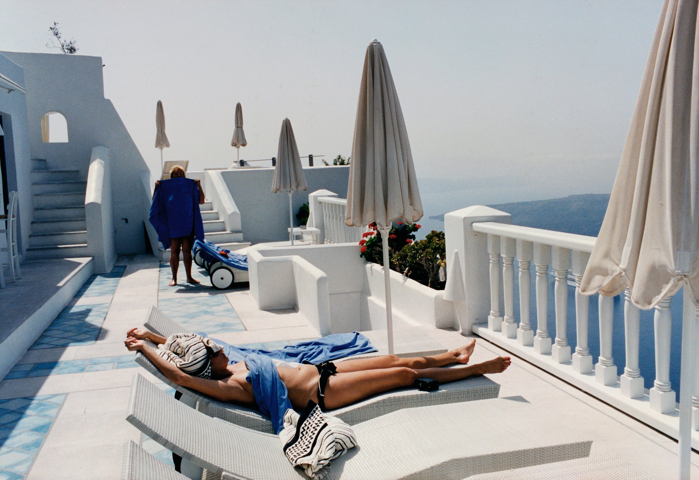 Woman sunbathing on a white chaise lounge on a rooftop patio with umbrellas, set against a blue seascape and clear sky.