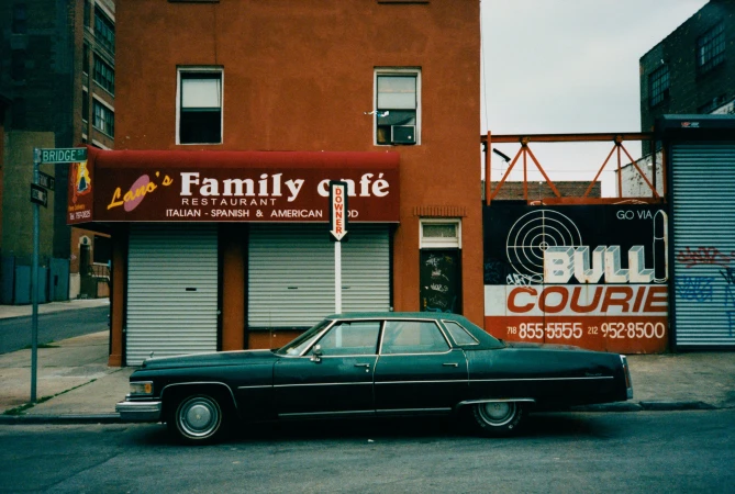 Classic dark green sedan parked outside Lano's Family Cafe restaurant. Building with Bull Courie sign in urban street scene.