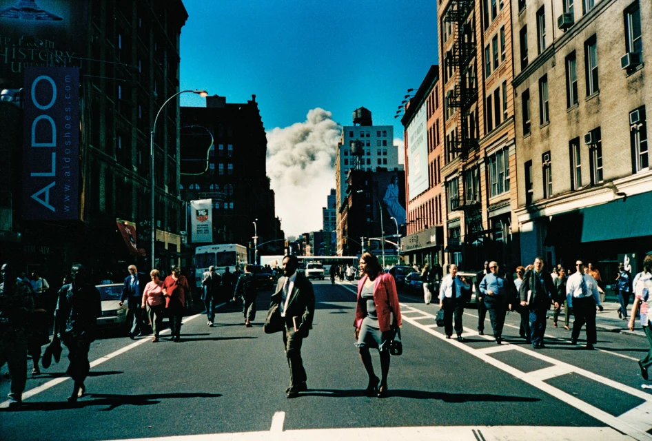 City street scene with pedestrians crossing road, buildings lining the sides, and a smoke cloud in background against a blue sky.
