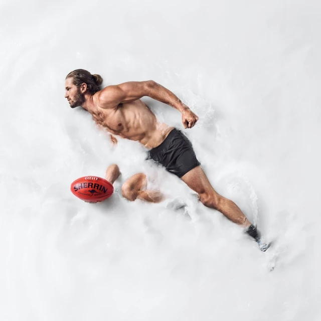 Athletic man running in white powder, wearing shorts and barefoot, with a red Sherrin Australian rules football to the side. 