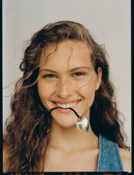 Close-up portrait of a smiling woman with long brown curly hair holding a silver heart pendant in her teeth.