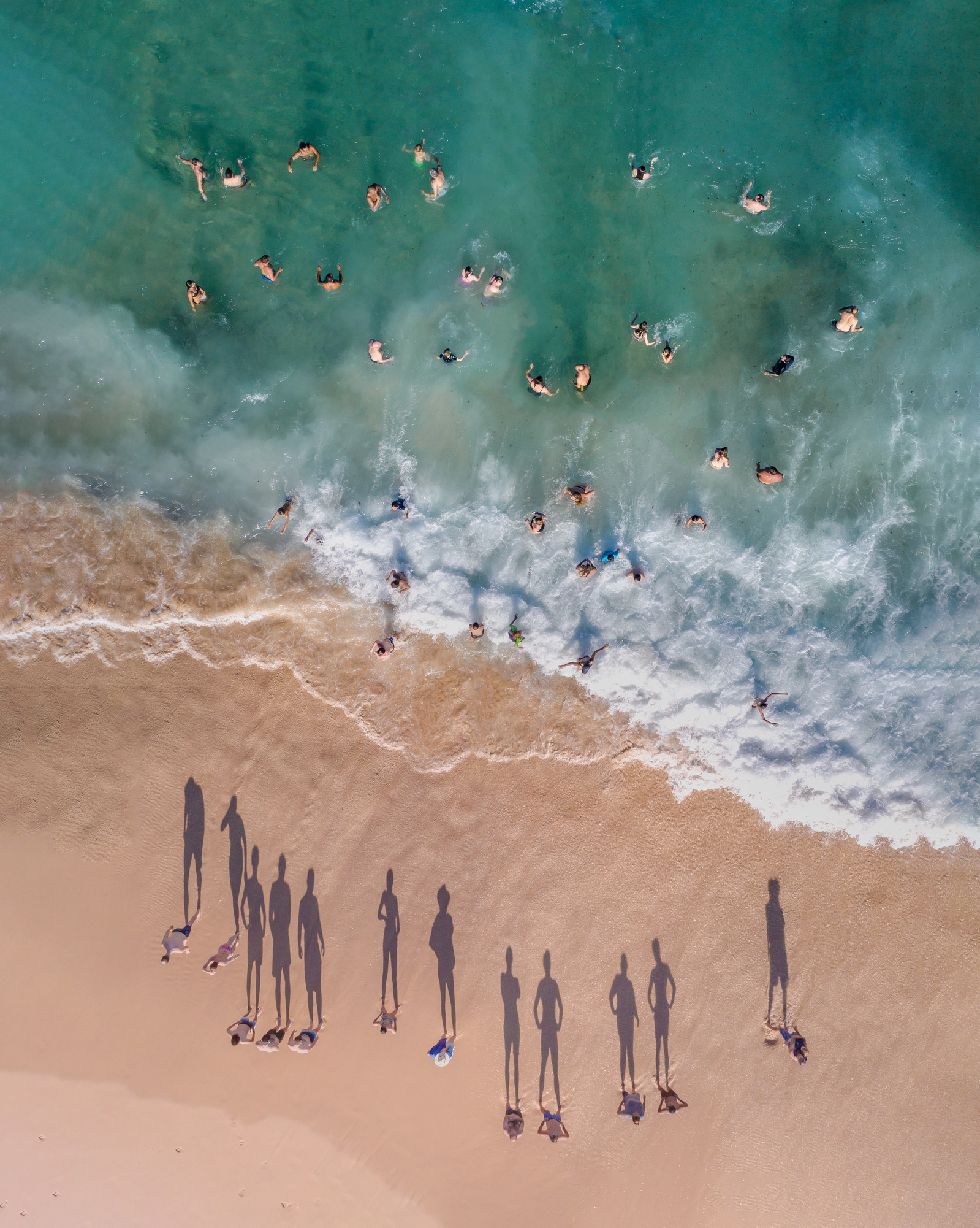 Aerial view of a sandy beach with groups of people standing on the sand and swimming in the ocean with turquoise water and foamy waves.
