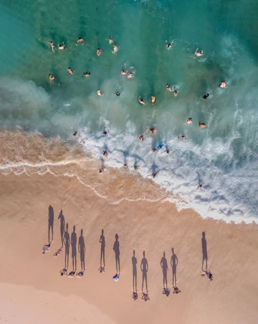 Aerial view of a sandy beach with groups of people standing on the sand and swimming in the ocean with turquoise water and foamy waves.