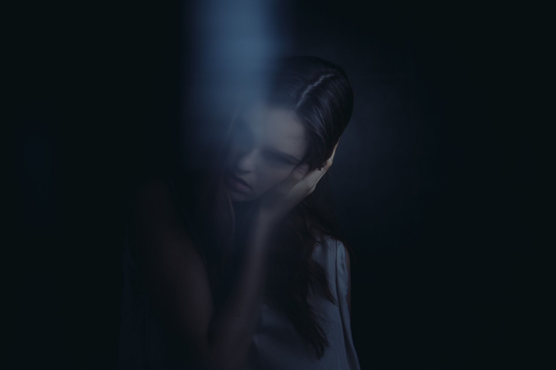 Portrait of a woman with dark hair and pale skin, holding her hand to her face, set against a dark, moody background with soft lighting.