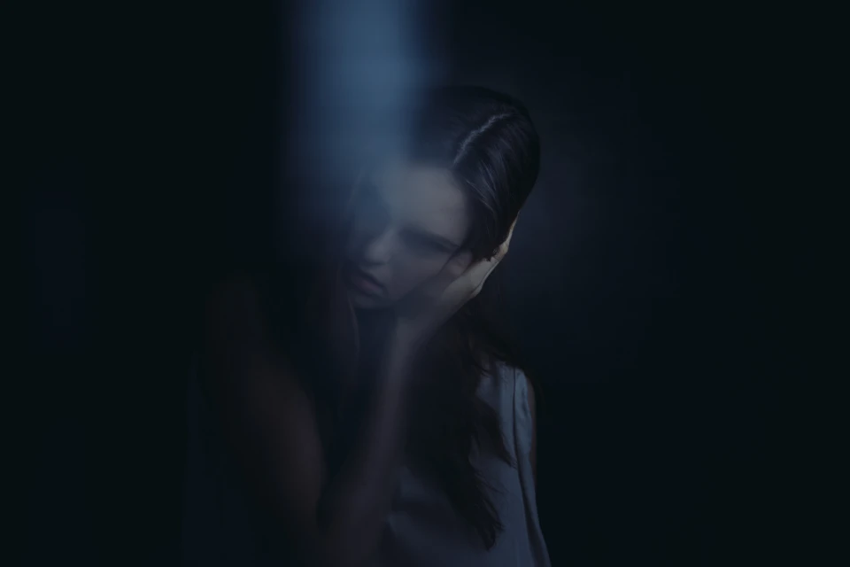 Portrait of a woman with dark hair and pale skin, holding her hand to her face, set against a dark, moody background with soft lighting.