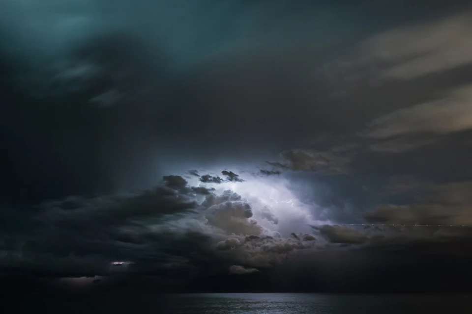 Dramatic nightscape featuring lightning illuminating dark clouds over a calm body of water, creating a powerful contrast of light and shadow.