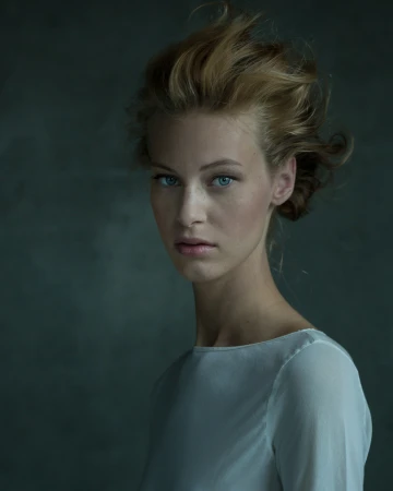 Portrait of a woman with blonde hair and blue eyes wearing a white top against a textured dark background by Irenaeus Herok.