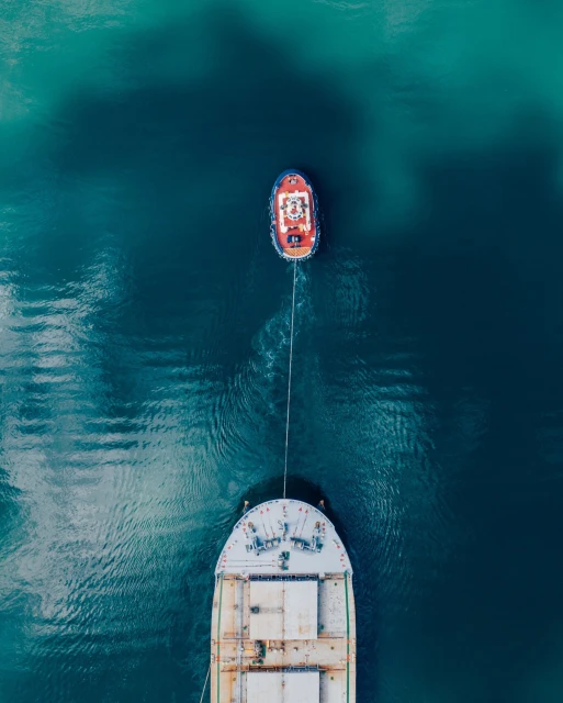 Aerial view of a tugboat towing a large cargo ship in teal water, showcasing the scale and dynamics of maritime transport.