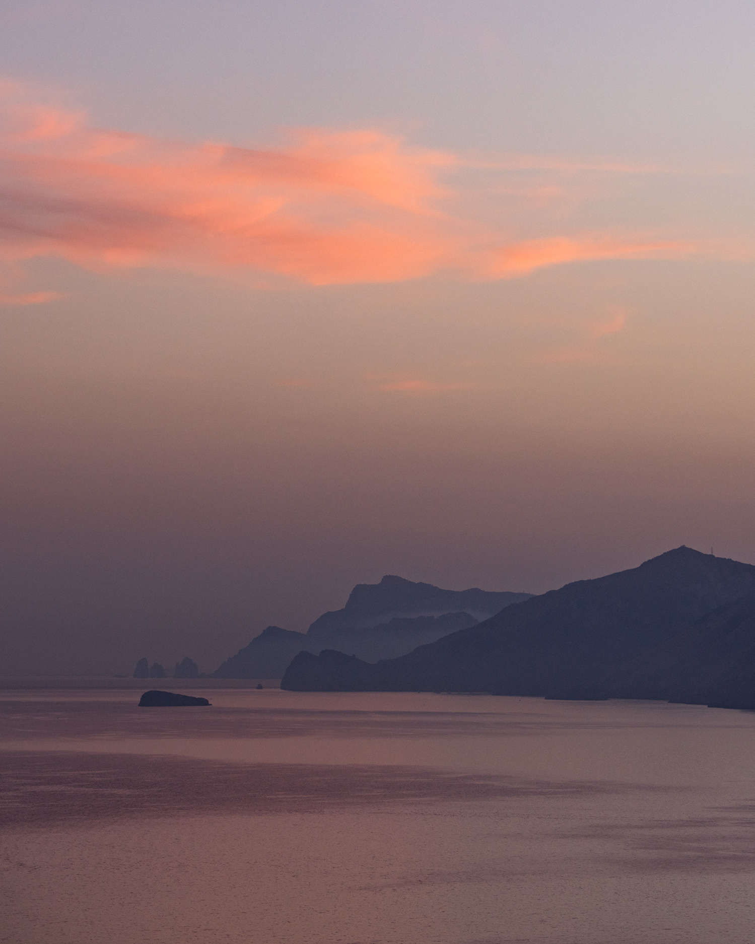 Scenic view of the ocean under a soft pastel sky at dusk, featuring distant mountains and the calm, reflective surface of the water.