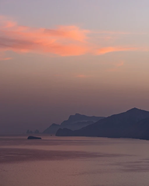 Scenic view of the ocean under a soft pastel sky at dusk, featuring distant mountains and the calm, reflective surface of the water.