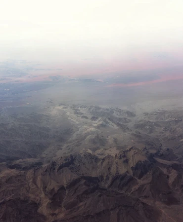 Aerial view of a rugged mountain range with dusty, arid terrain and a hazy sky, showcasing a remote and desolate landscape.