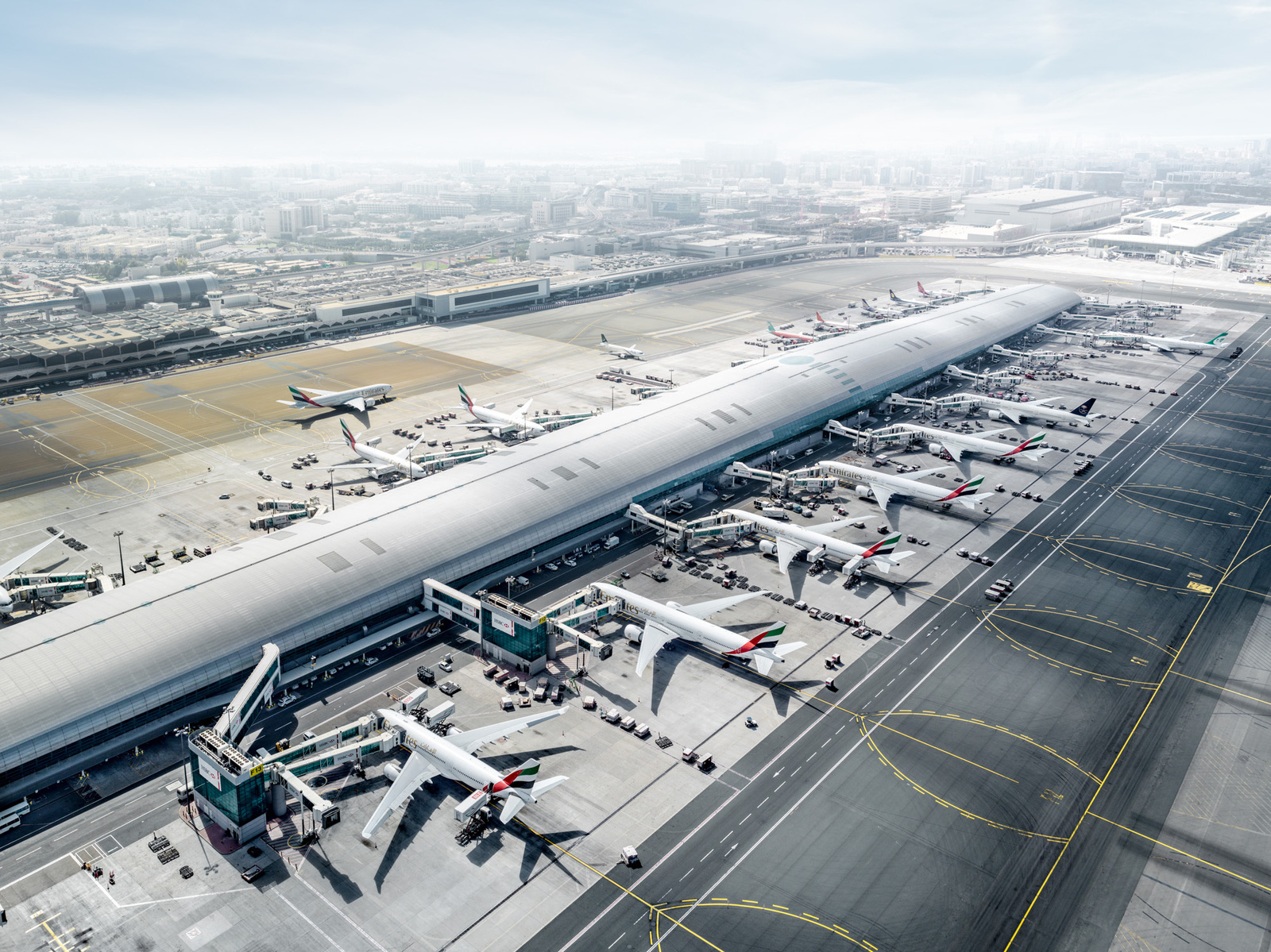 Aerial view of a modern airport terminal with multiple airplanes at the gates, featuring a cityscape in the hazy background on a sunny day.