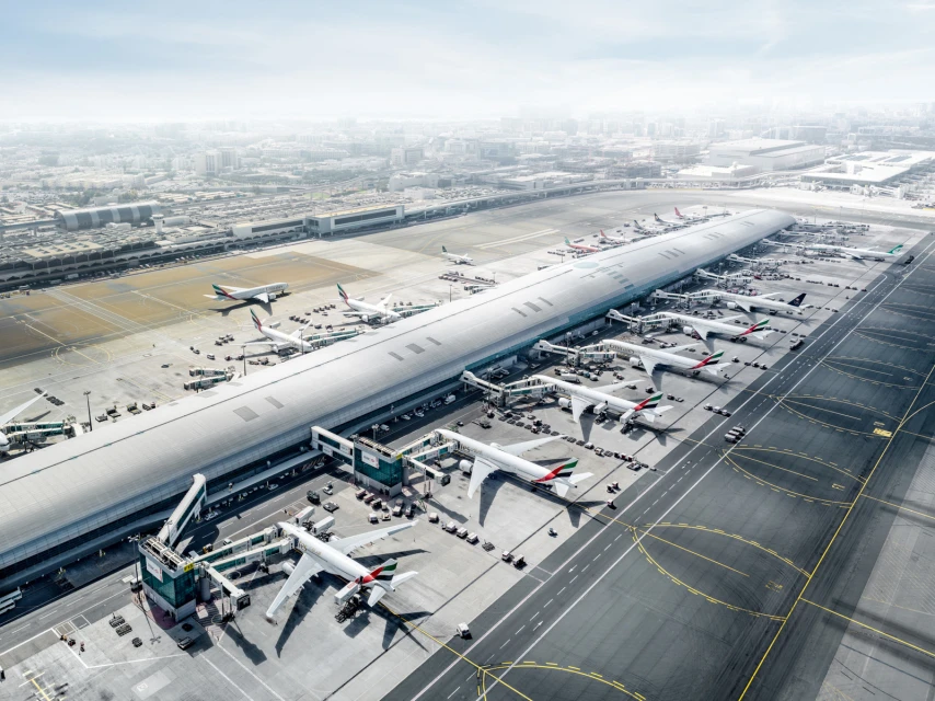 Aerial view of a modern airport terminal with multiple airplanes at the gates, featuring a cityscape in the hazy background on a sunny day.