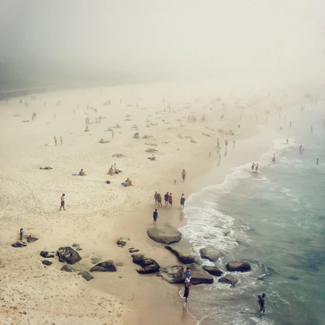 Sandy beach scene with many people, waves washing ashore, rocks in the foreground, and a soft, diffused light due to fog.