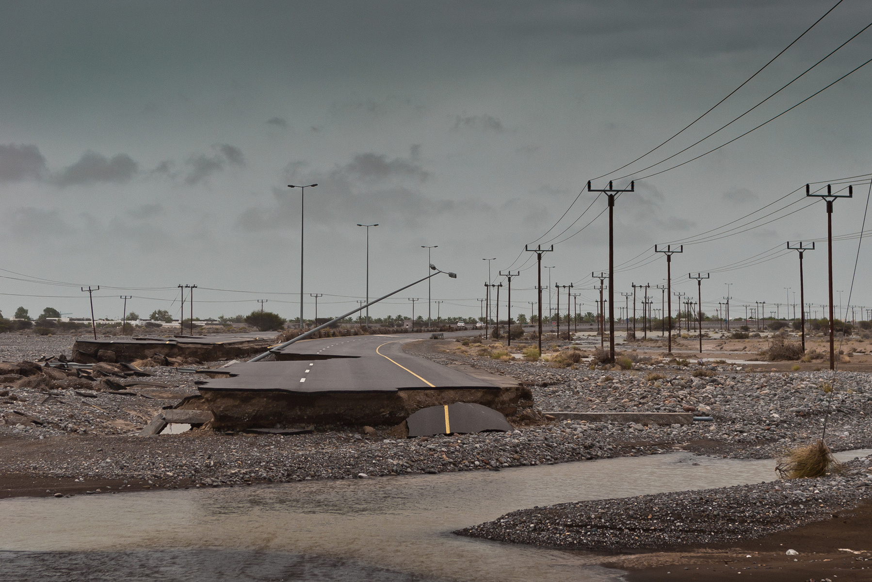 Damaged asphalt road with eroded edges runs through a rocky riverbed under an overcast sky with utility poles lining the background.