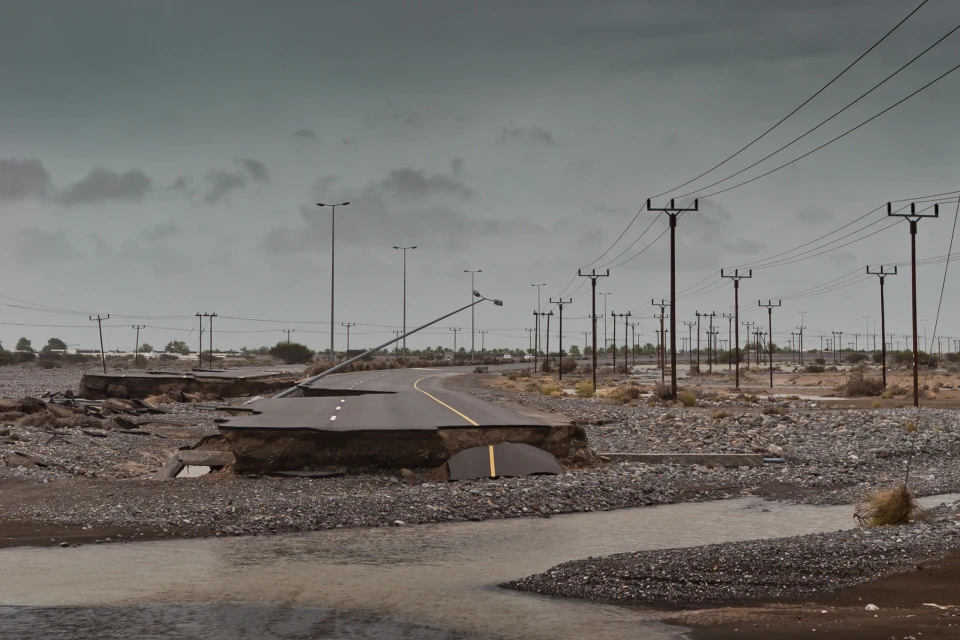 Damaged asphalt road with eroded edges runs through a rocky riverbed under an overcast sky with utility poles lining the background.