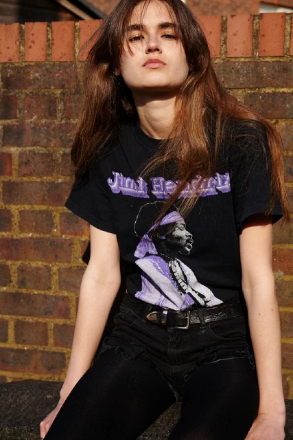 Portrait of a woman with long brown hair wearing a Jimi Hendrix t-shirt, posing in front of a brick wall in natural outdoor light.