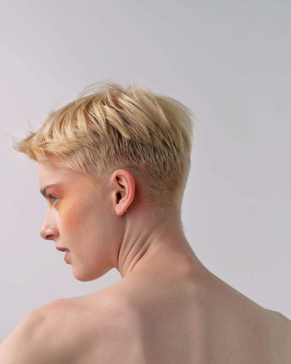 Close-up studio shot of a fair-skinned adult female with a blonde short haircut, bare shoulders, and colourful eyeshadow on a neutral background.