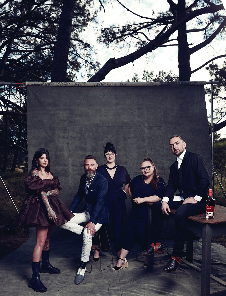 Group portrait of five adults with varied hairstyles and fashion in an outdoor studio setting against a grey backdrop with forest trees.