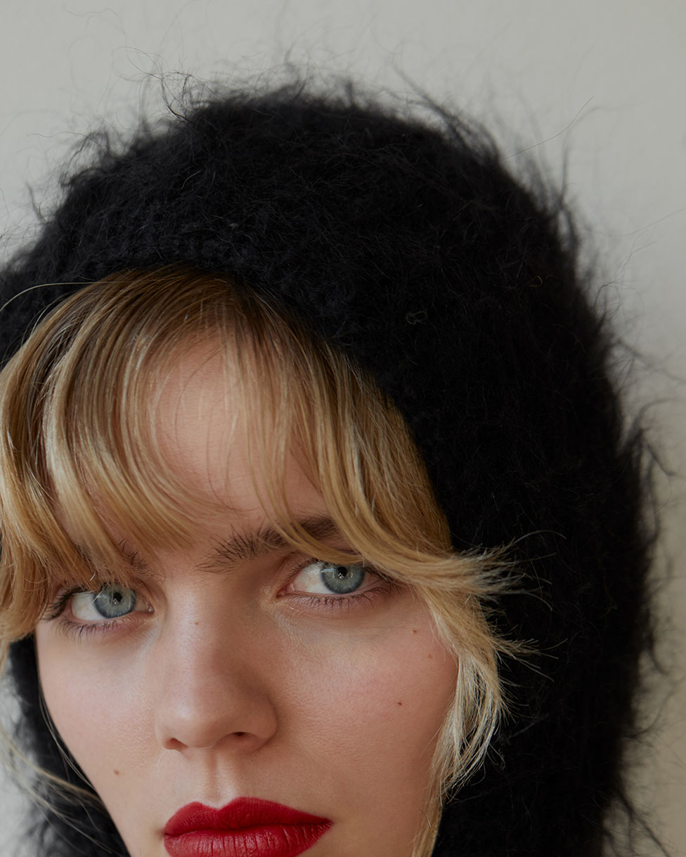 Close-up of a blonde woman with blue eyes and red lipstick wearing a fuzzy black head covering against an off-white background. Fashion shot.