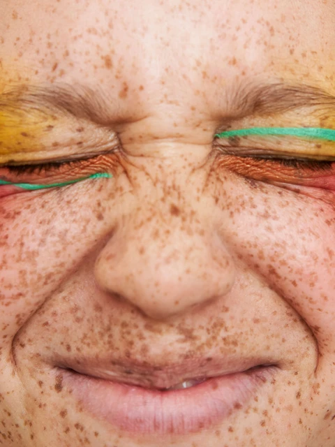 Close-up of a fair-skinned adult female's face with freckles, closed eyes adorned with colorful makeup, including yellow, copper, and green.