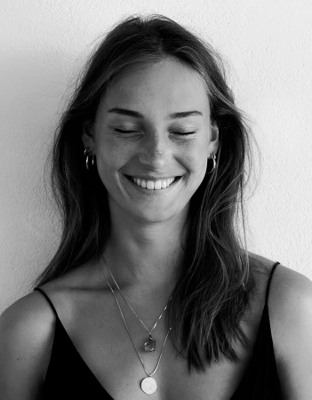 Portrait of a smiling woman with freckles and long dark hair wearing delicate layered necklaces and hoop earrings against a white wall.