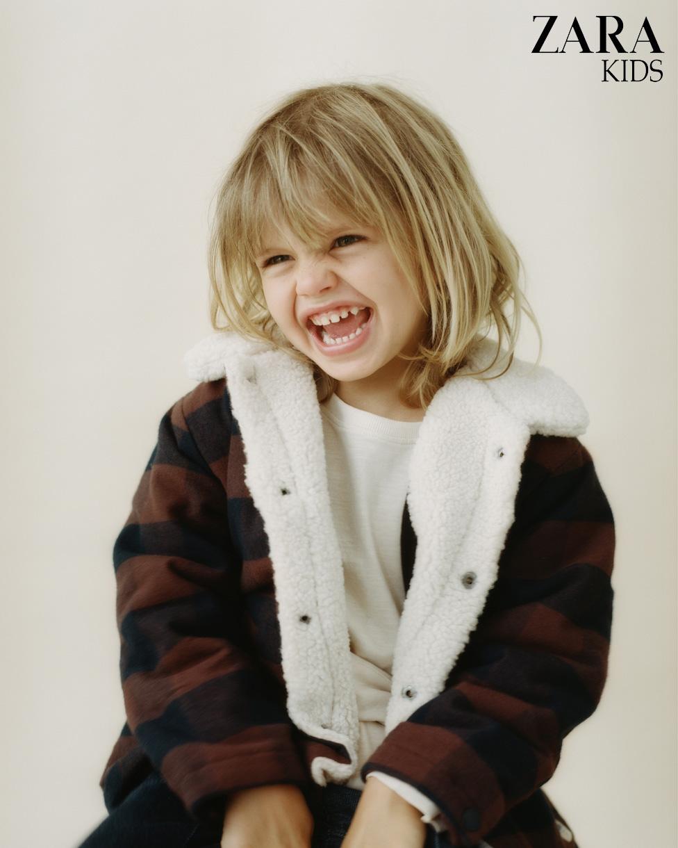 Child model grinning in a Zara Kids ad, wearing a plaid coat with a sherpa collar against a light background. Zara Kids logo in corner.