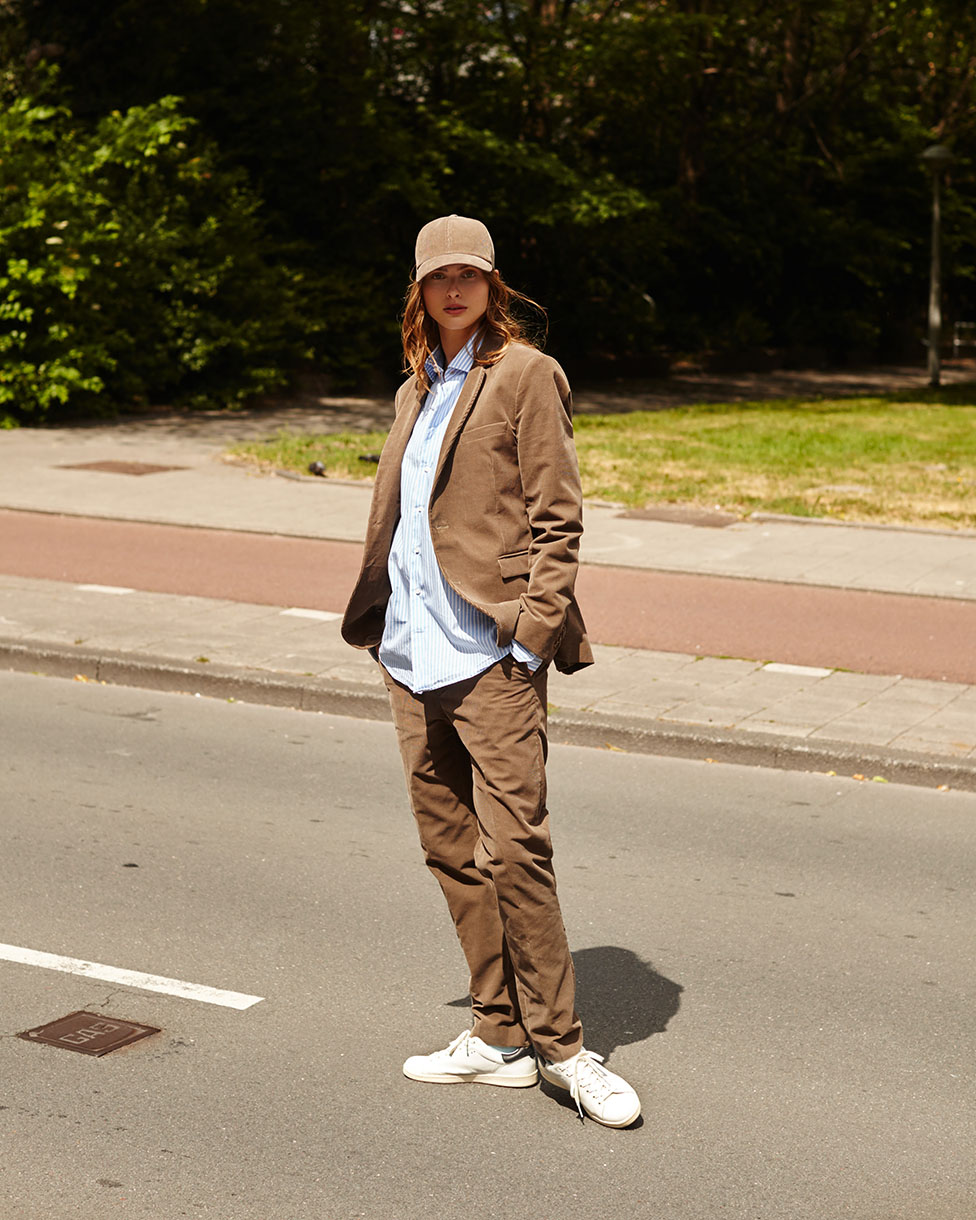 Woman in a brown suit and baseball cap stands on a sunny city street; trees in the background. Casual yet stylish fashion.