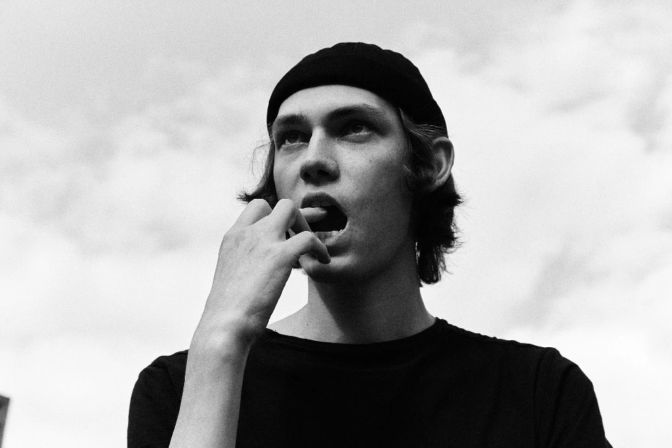 Low angle black and white portrait of a young adult man with a beanie looking up with his tongue out against a cloudy sky.