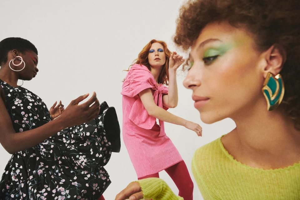 Three diverse women in stylish dresses and vibrant makeup pose against a clean white backdrop in a fashion-forward studio shot.