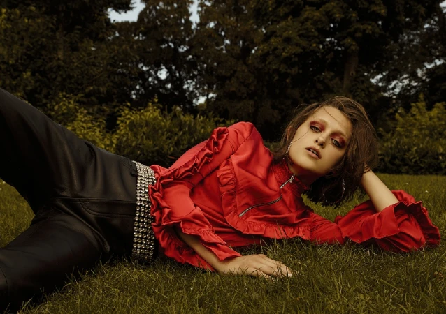 Model lying on grass wearing a red ruffled blouse, dark pants, and a jeweled belt, styled by Charlotte Niketic.
