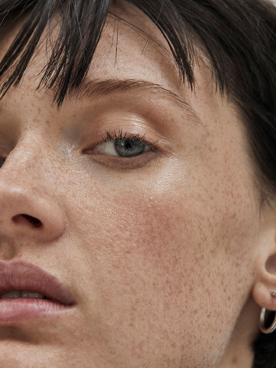 Extreme close-up of a young woman's face showcasing natural skin texture, freckles, dark hair, blue eyes, and small hoop earrings.