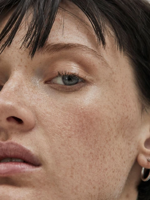 Extreme close-up of a young woman's face showcasing natural skin texture, freckles, dark hair, blue eyes, and small hoop earrings.