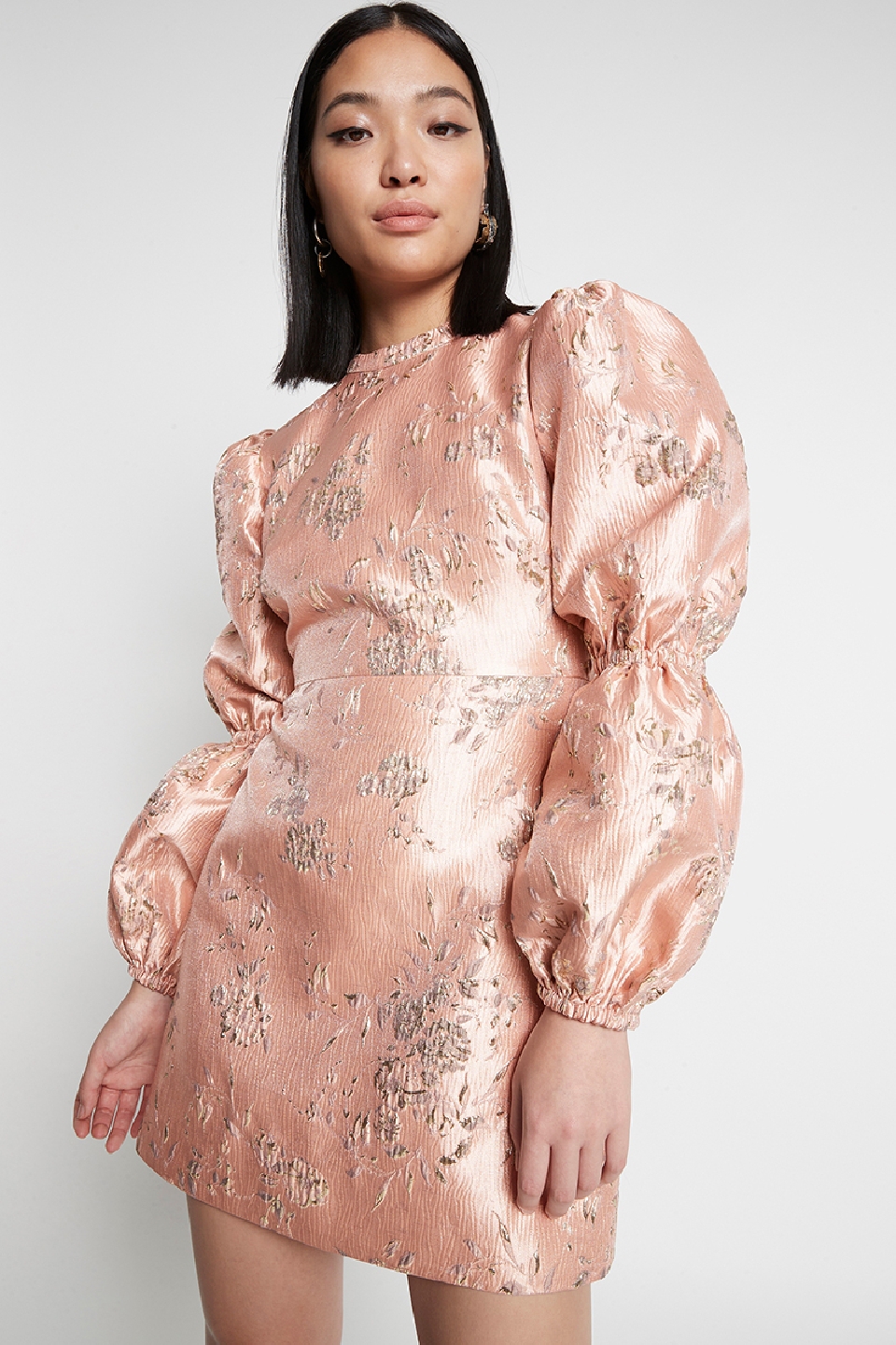 Woman in a peach-colored mini dress with floral details and puffy sleeves standing against a white background in a studio setting.