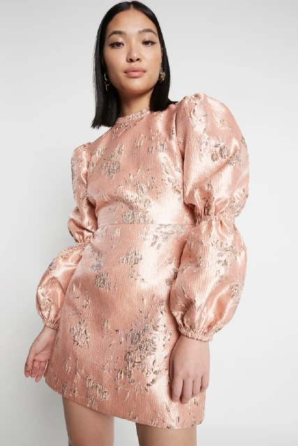 Woman in a peach-colored mini dress with floral details and puffy sleeves standing against a white background in a studio setting.