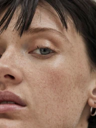 Close-up beauty portrait of a woman with freckles, short dark hair, and natural makeup featuring a silver hoop earring.