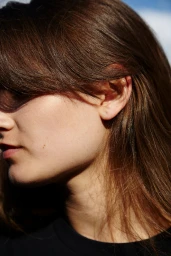 Close-up profile of a woman with long brown hair wearing a black top against a clear blue sky background.