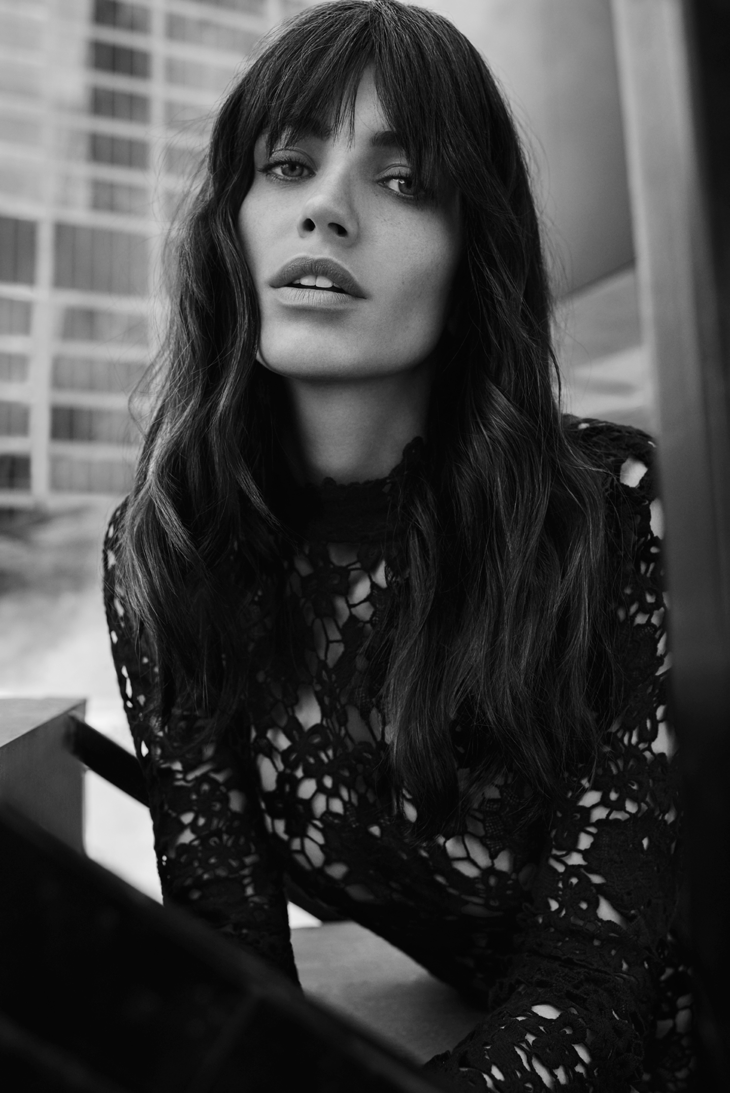 Black-and-white portrait of a woman with bangs, wearing a lace top, against a windowed background in an indoor environment.