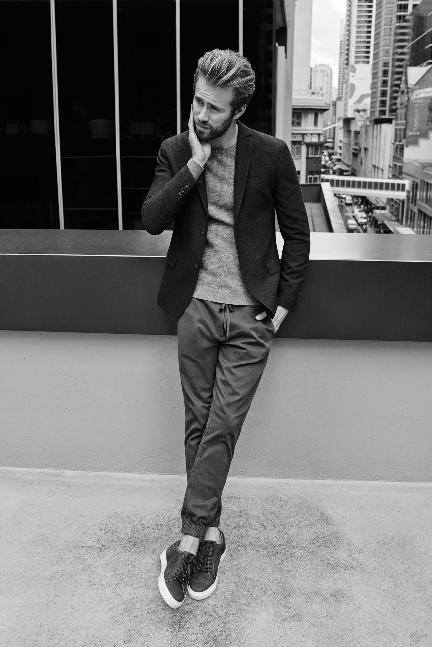 Man wearing a blazer, sweater, and trousers leaning against a balcony railing with city buildings in the background.