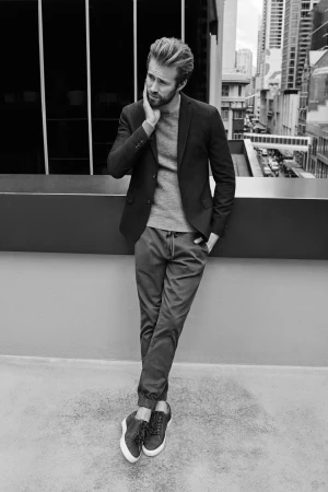 Man wearing a blazer, sweater, and trousers leaning against a balcony railing with city buildings in the background.