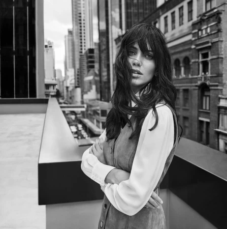 Monochrome portrait of a woman with long dark hair and bangs standing on a rooftop with city buildings in the background.