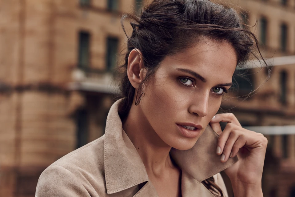 Close-up portrait of a young adult woman in a tan jacket posing outdoors with an urban building visible in the background.