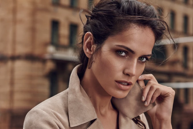 Close-up of a brunette adult female model with green eyes wearing a beige suede jacket, posing outdoors with a blurred building background.