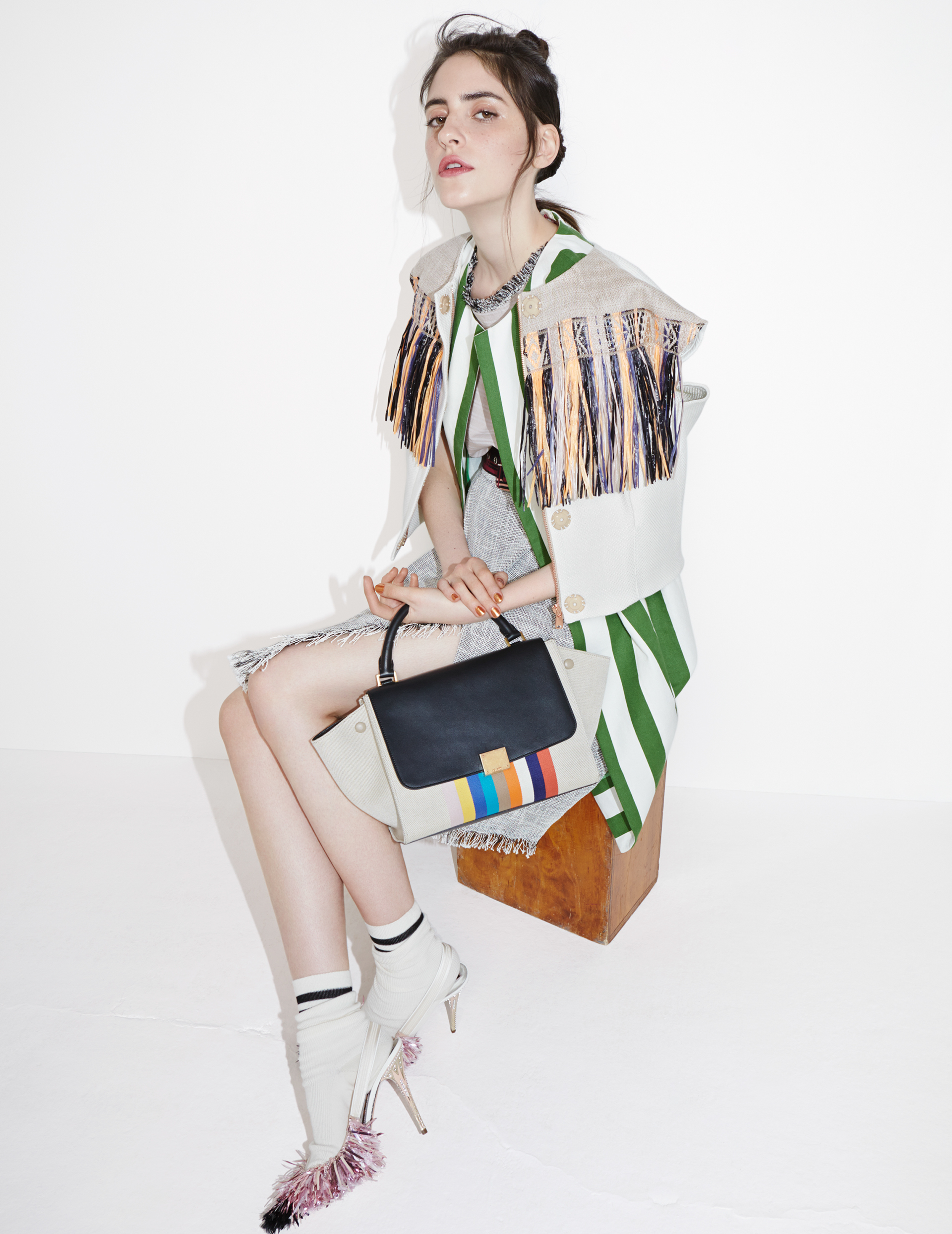 A woman in a striped jacket sits on a wooden stool, holding a color-blocked handbag, while wearing white high heel shoes with pink trim.