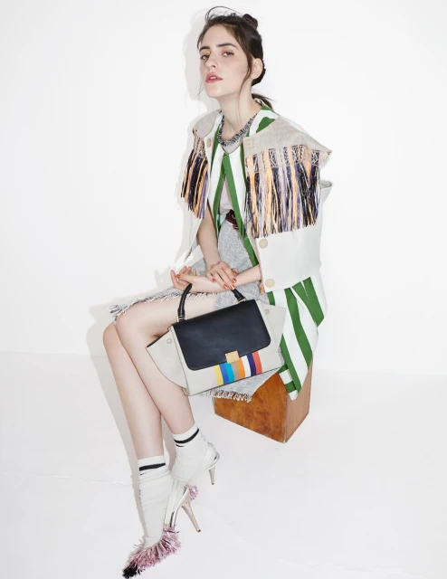 A woman in a striped jacket sits on a wooden stool, holding a color-blocked handbag, while wearing white high heel shoes with pink trim.